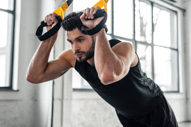 handsome concentrated sporty man hanging on resistance bands and looking away in gym - Φωτογραφία, εικόνα