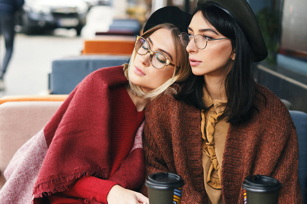 Autumn winter portrait of two fashionable women in an outdoor cafe, drinking coffee to go, posing. Urban background. Sisters - Photo, Image
