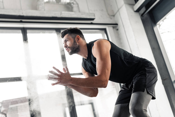 Tiefansicht eines athletischen jungen Mannes, der Talkumpuder auf die Hände aufträgt und im Fitnessstudio wegschaut - Foto, Bild