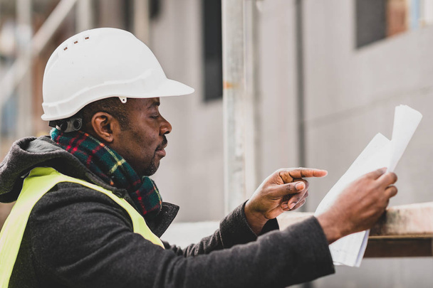 Ingeniero masculino afroamericano absorbido con chaqueta de seguridad y casco comprobando dibujos técnicos y planos de oficina entre andamios
 - Foto, Imagen