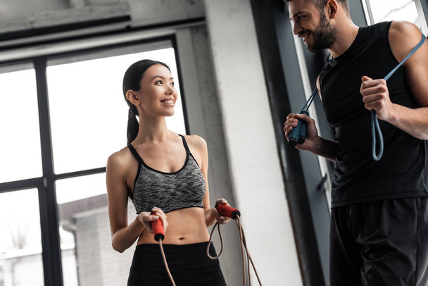 happy sporty young couple holding jumping ropes and smiling each other in gym - Foto, Imagem
