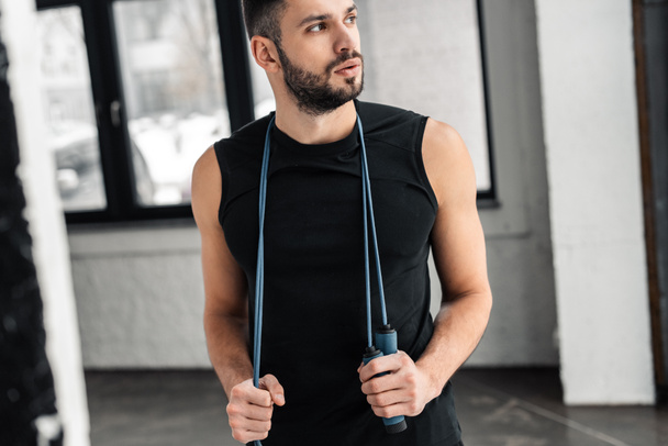 cropped shot of handsome sportsman holding skipping rope around neck and looking away in gym  - Zdjęcie, obraz