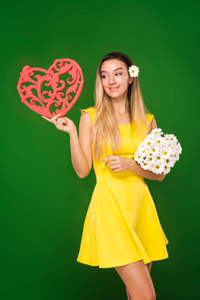 mooi blonde meisje in een gele jurk heeft een hart in haar handen en een boeket van chamomiles  - Foto, afbeelding
