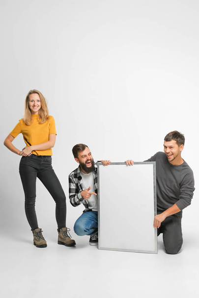 three funny laughing young people wearing casual fashionable clothes, on hunkers, posing, holding mockup big white board isolated in studio, white background - Foto, afbeelding