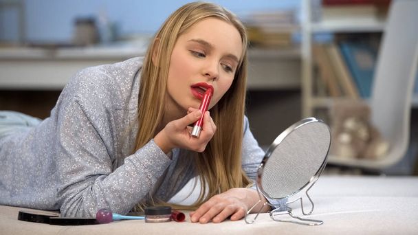Teenager girl applying red lipstick, looking in mirror, getting ready to go out - 写真・画像