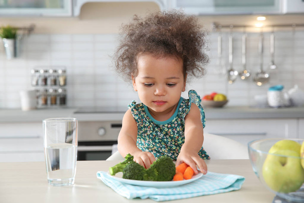Cute African-American girl eating vegetables at table in kitchen - Zdjęcie, obraz