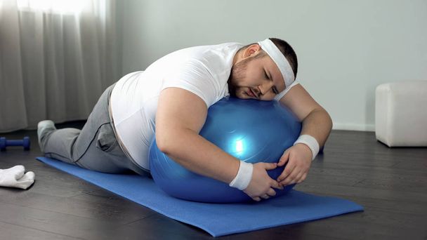 Weak-willed fat man relaxing on fitness ball, home workout break, laziness - Valokuva, kuva