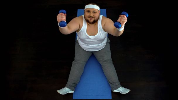 Active fat man lifting dumbbells sitting on mat, sport motivation, discipline - Fotografie, Obrázek
