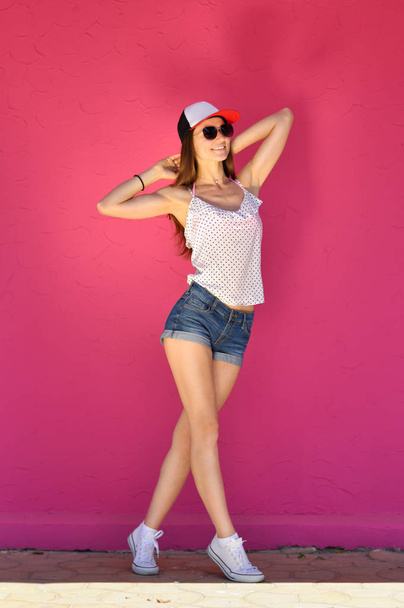 Hip hop tarzı giyinmiş, güneş gözlüklü seksi genç kadın pembe duvar arkasında poz veriyor. - Fotoğraf, Görsel