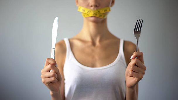 Anorexic girl holding fork and knife, mouth closed with tape, restrictions - Foto, imagen
