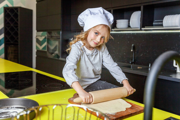 Little girl with chef hat, rolling pin preparing dough, bake homemade holiday apple pie in kitchen. Kid cooking healthy food at home and having fun. Childhood, household, teamwork helping concept - Photo, image