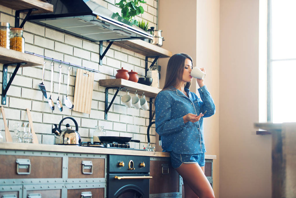Attractive beautiful woman in blue pajamas is drinking tea or coffee at home in kitchen in the morning. - Foto, afbeelding