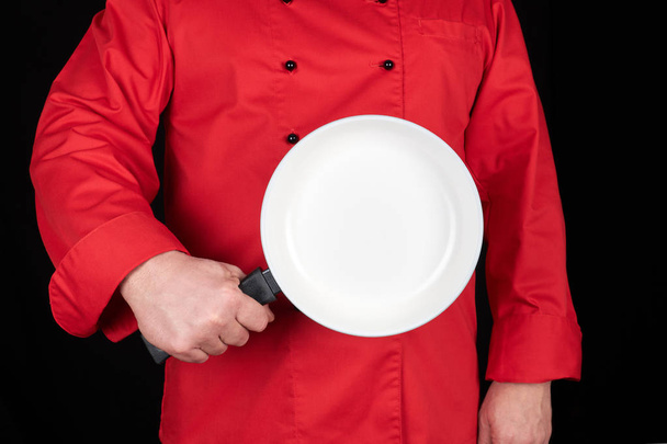 cook in red uniform holding an empty round white frying pan, black background - Zdjęcie, obraz