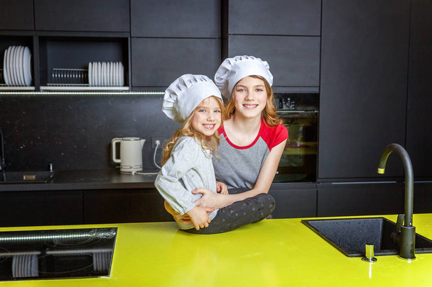 zwei Mädchen mit Kochmütze, die sich umarmen und Spaß in der Küche haben. Schwestern kleines Kind und Teenager Mädchen kochen gesunde Lebensmittel zu Hause. Kindheit, Familie, Haushalt, Teamwork-Helferkonzept - Foto, Bild