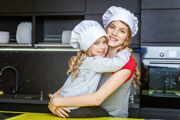 Dwie dziewczyny z szefa kuchni kapelusz, przytulanie i zabawy w kuchni. Siostry mały dzieciak i nastolatkę zdrowego gotowania w domu. Dzieciństwo, rodziny, gospodarstwa domowego, pracy zespołowej w pomagając koncepcja - Zdjęcie, obraz