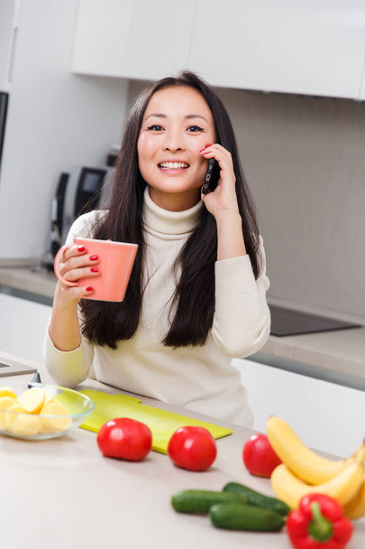 Image of young brunette with mug and phone in hands standing at table with vegetables and fruits - Photo, Image