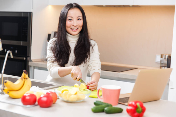 Foto de joven asiática chica con taza y portátil de pie en la mesa con verduras y frutas
 - Foto, Imagen