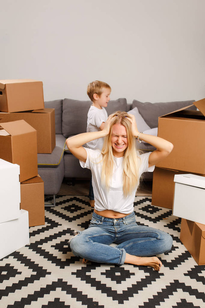 Photo of upset blonde woman sitting on floor among cardboard boxes and boy near gray sofa - Foto, afbeelding