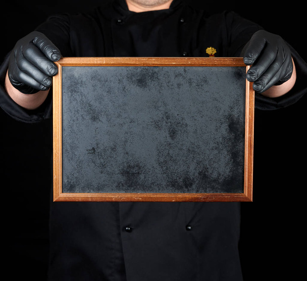 σεφ στην μαύρη στολή και μαύρα γάντια λατέξ κατέχει μια κενή ξύλινη κορνίζα, αντίγραφο χώρου - Φωτογραφία, εικόνα