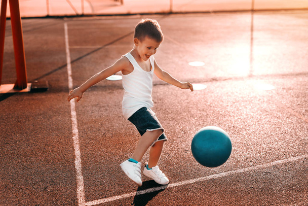 Petit garçon en short de sable chemise donnant des coups de pied au ballon sur le court le matin
. - Photo, image