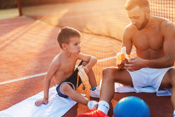 Shirtless father and son sitting on the court, eating bananas and resting after exercising. Summertime at morning. - Foto, afbeelding