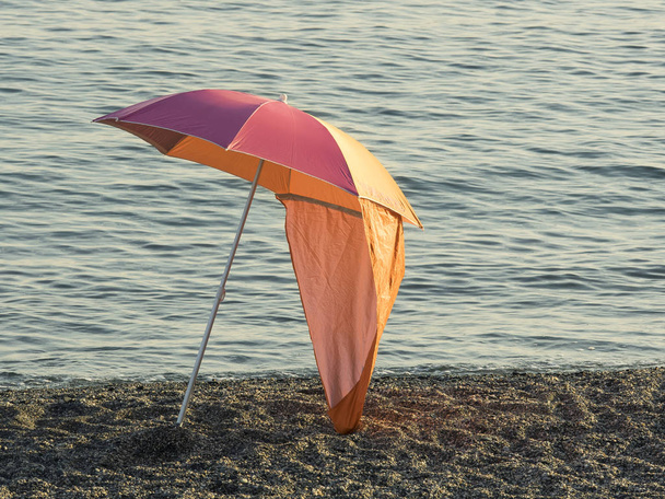 bunter Sonnenschirm (Sonnenschirm) isoliert auf einem Kiesstrand bei Sonnenuntergang ruhiges Meerwasser im Hintergrund - Foto, Bild