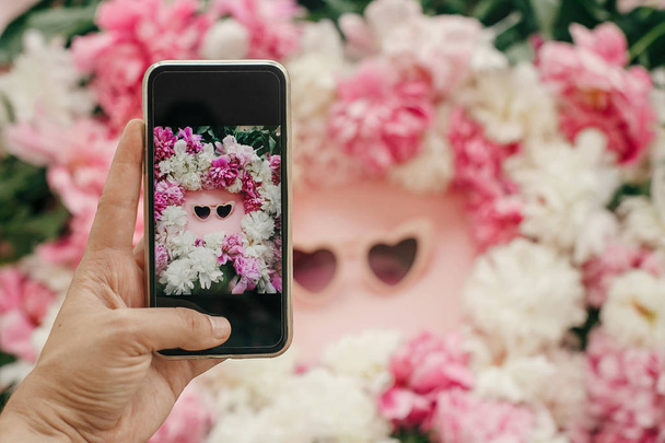 Instagram blogging concept. Hand met telefoon en nemen foto van stijlvolle girly flat leg hart vorm roze zonnebril met roze en witte pioenrozen op pastel roze papier. Hallo zomer - Foto, afbeelding