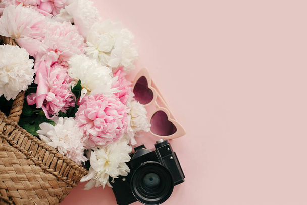 Stylish  pink peonies in straw bag, photo camera, retro sunglasses on pastel pink paper with copy space. International Women's Day. Hello spring flat lay. Summer vacation concept - Photo, Image
