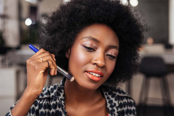 Pretty african american woman with curly hair putting a new concealer on her face - Photo, image