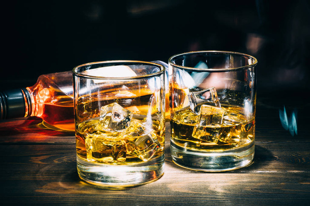 Dvě sklenky skotské whisky nebo koňaku s kostkami ledu a láhev alkoholu alkohol na tmavé dřevěné pozadí - Fotografie, Obrázek