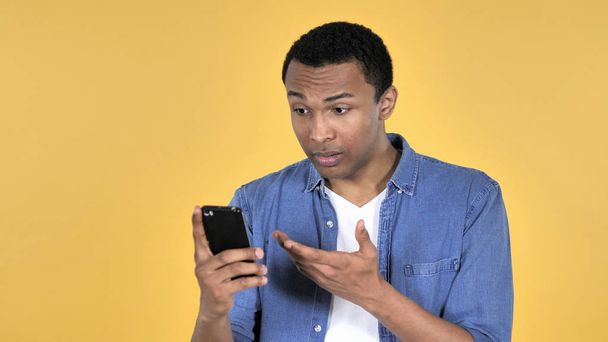 Jonge Afrikaanse Man in Shock tijdens het gebruik van Smartphone geïsoleerd op gele achtergrond - Foto, afbeelding