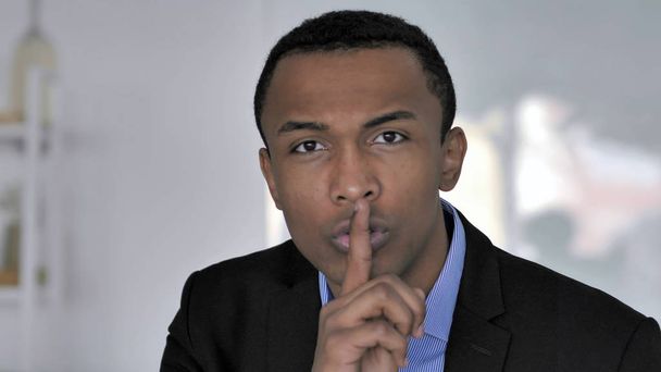 Porträt eines afrikanischen Geschäftsmannes, der mit dem Finger auf den Lippen Schweigen gestikuliert - Foto, Bild