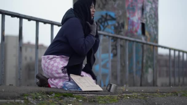 Elderly homeless woman with outstretched hand sits on the bridge in cold windy grey weather asking for alms and help and one woman stopped to put coins - Felvétel, videó