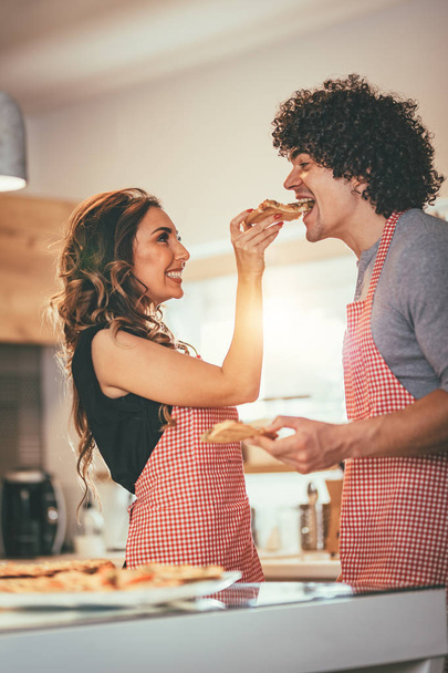 Nuori pari tekee ja maistaa pizzaa keittiössä
 - Valokuva, kuva