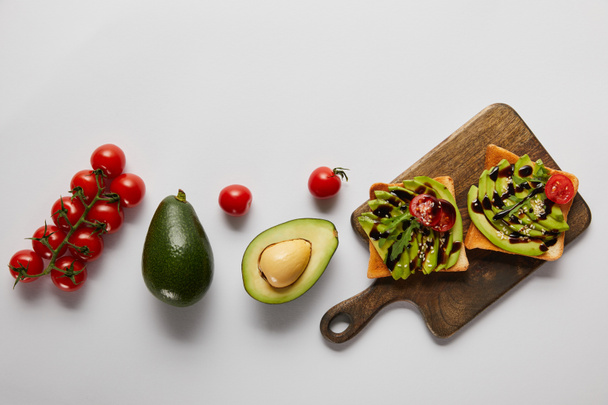 top view of toasts on wooden cutting board with avocados and cherry tomatoes on grey backgroud - Photo, Image