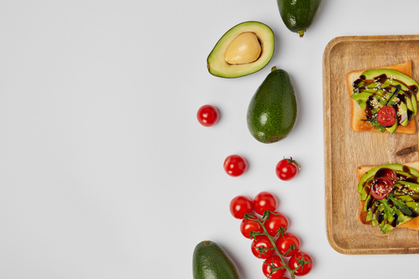 top view of toasts on wooden cutting board with avocados and cherry tomatoes on grey backgroud - Fotoğraf, Görsel