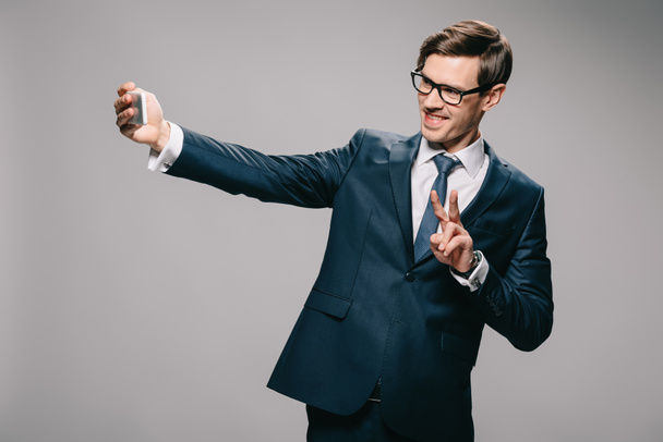 empresário alegre tomando selfie e mostrando sinal de peça no fundo cinza
  - Foto, Imagem