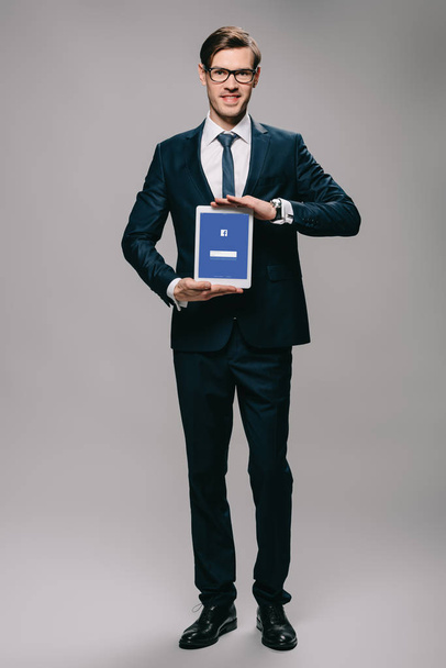 handsome businessman holding digital tablet with facebook app on screen on grey background  - Foto, afbeelding
