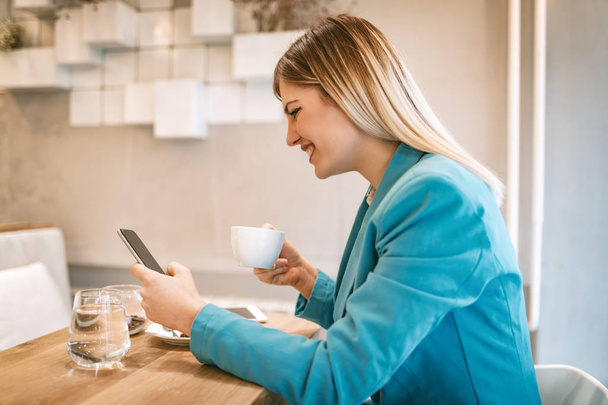 Молодая улыбающаяся деловая женщина на кофе-брейке в кафе с помощью смартфона
  - Фото, изображение
