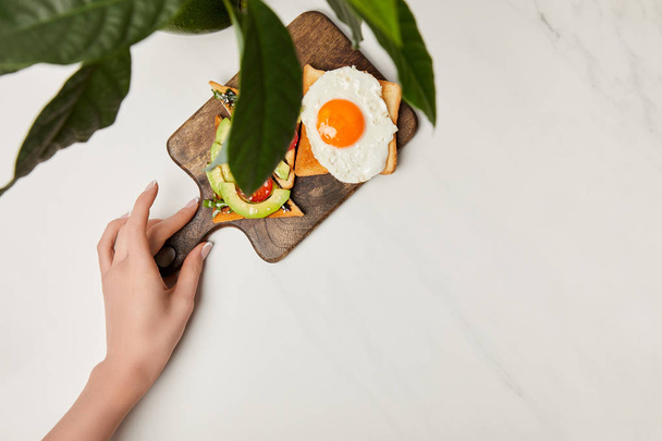 top view of womans hand holding wooden cutting board with toasts and scrambled egg under green plant on marble surface - Photo, Image