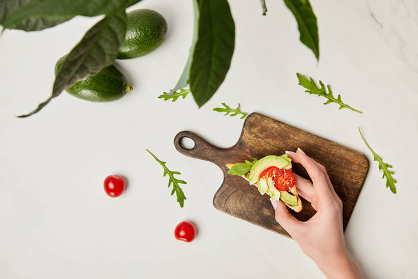top view of womans hand holding toast, wooden cutting board, avocados and cherry tomatoes under green plant on marble surface - Photo, Image
