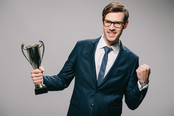 handsome businessman in glasses celebrating victory and holding trophy in hand on grey background - Photo, Image