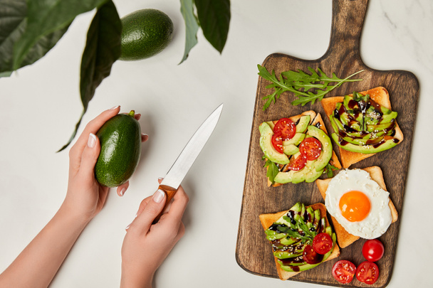 top view of womans hands holding knife with avocado and wooden cutting board with toasts on marble surface - Foto, immagini