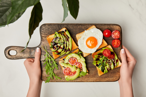 top view of womans hands holding wooden cutting board with toasts and scrambled egg under green plant on marble surface - Foto, Imagem