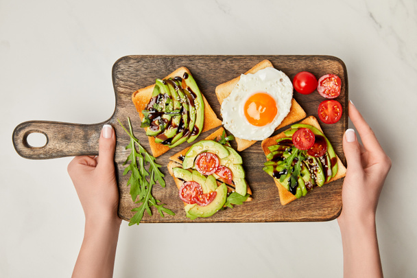 top view of womans hands holding wooden cutting board with toasts and scrambled egg on marble surface - Photo, Image