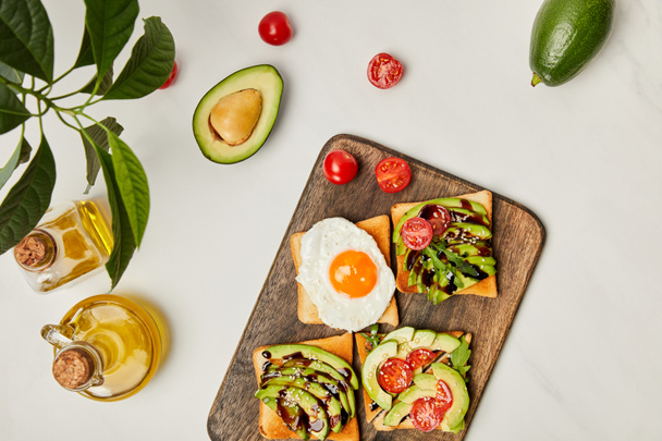 top view of wooden cutting board with toasts, scrambled egg, cherry tomatoes, avocados and oil bottles on marble surface - Foto, afbeelding