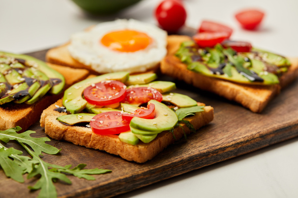 selective focus of cutting board with toasts, scrambled egg, cherry tomatoes and avocado on white background - 写真・画像