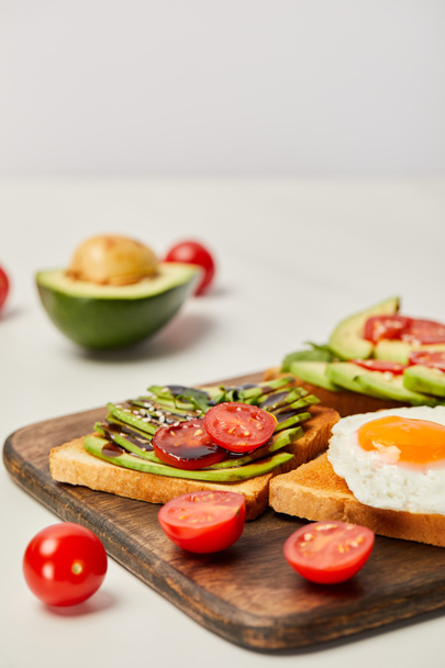 Selektivní fokus na dřevěném prkénku s toasty, míchaná vejce, cherry rajčaty a avokádem na šedém pozadí - Fotografie, Obrázek