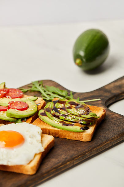 selective focus of wooden cutting board with toasts, scrambled egg, cherry tomatoes and avocado on grey background - Photo, Image