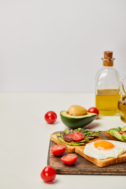 Ahşap kesme tahtası tost, çırpılmış yumurta, domates, avokado ve gri arka plan üzerinde yağ şişeleri ile seçici odak - Fotoğraf, Görsel